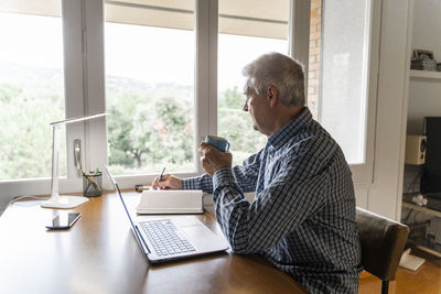 Senior man sitting at home in front of laptop making notes