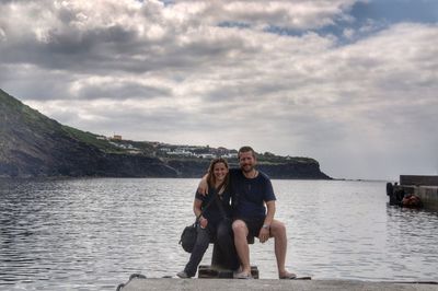 Portrait of couple sitting on wooden stump by lake against cloudy sky