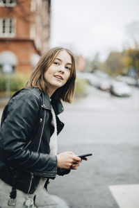Smiling girl with mobile phone looking away while standing on street in city