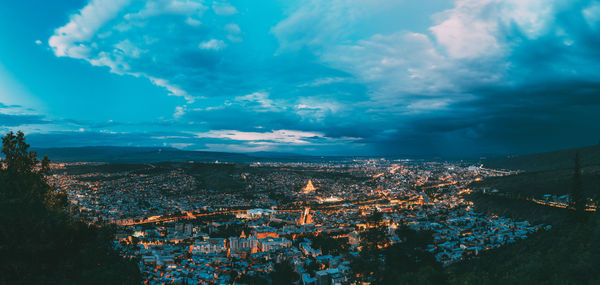 Aerial view of illuminated buildings in town against cloudy sky