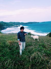Man checking time while standing by sheep on grass against sea