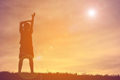 Silhouette boy with hand raised standing on field against cloudy sky during sunset