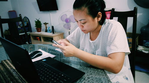 Young woman using phone in front of laptop on table at home