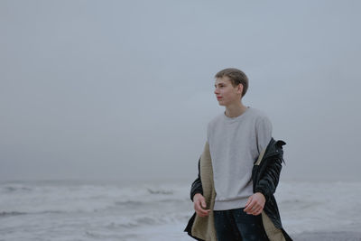 A teenager in a white sweatshirt stands against the background of the cloudy sea
