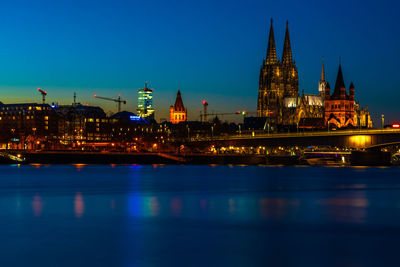 Illuminated buildings at waterfront against blue sky