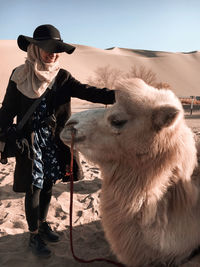 Woman wearing hat standing beside camel on sand dunes