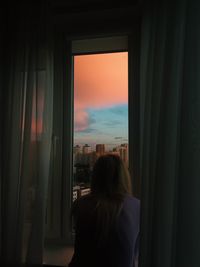 Rear view of woman looking through window at home during sunset