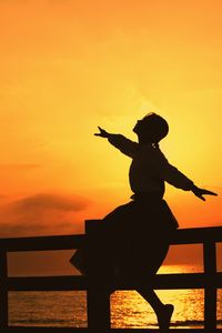 Silhouette woman with arms outstretched sitting on railing against orange sky during sunset