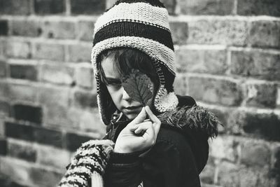 Boy in warm clothing holding leaf against brick wall