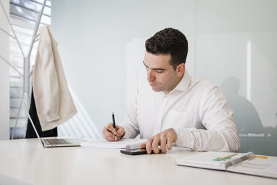 Young businessman with writing in book sitting at desk in office