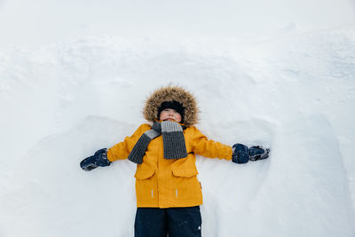 Cute little boy in an orange jacket with a fur hood is having fun lying in the snow. 