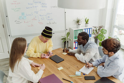 Businessman writing strategy on adhesive note sitting by colleagues in office