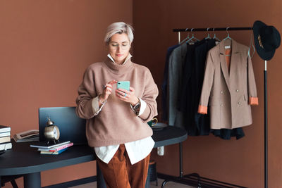 Fashion woman in brown oversize sweater and trousers standing in modern work place with smartphone
