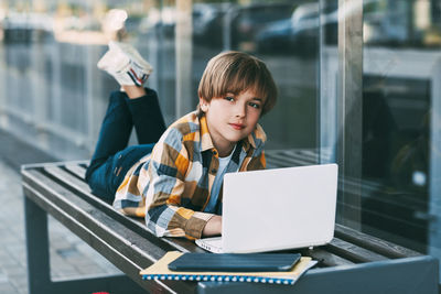 Boy using laptop while lying on bench by modern building