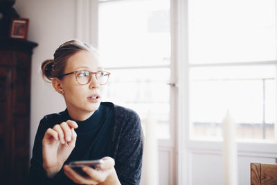 Young woman looking away while holding mobile phone against window at home