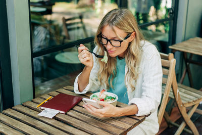 A young woman is sitting on the terrace in a cafe eating a salad and watching a video on her phone.