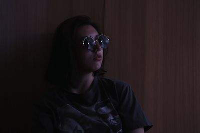 Contemplating woman looking away while wearing sunglasses against wooden wall