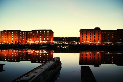 Illuminated city by river against sky at sunset