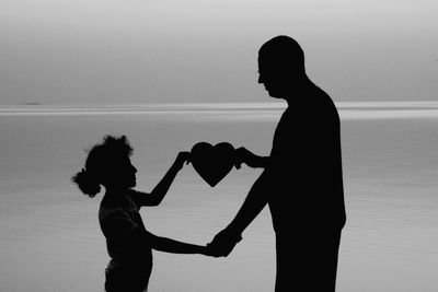 Silhouette father and daughter holding heart shape against sea