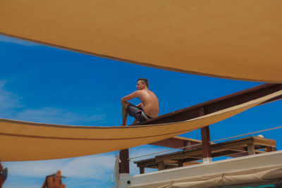 Low angle view of shirtless man sitting on pier against blue sky