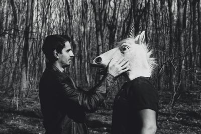 Side view of man standing with male friend wearing horse mask in forest