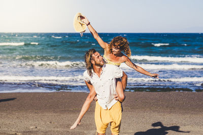 Man piggybacking cheerful woman at beach against sky