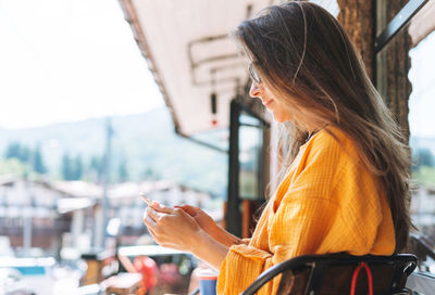 Young woman with long hair sunglasses and yellow longsleeve with mobile phone in hands in cafe