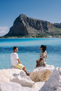 Friends sitting on rock by sea against mountain