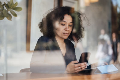 Woman using smart phone seen through glass at cafe