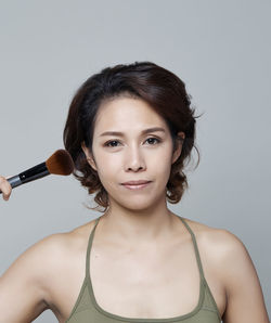 Portrait of smiling beautiful woman holding make-up brush against gray background
