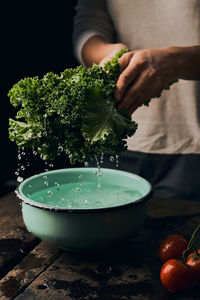 Unrecognizable crop housewife washing green curly kale in bowl with water at wooden table on black background