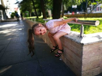 Side view of playful girl sitting on retaining wall by footpath