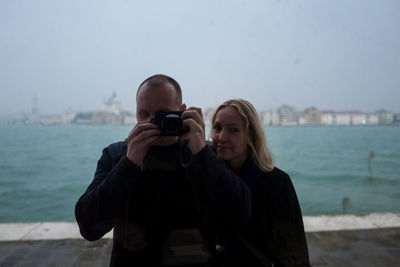 Portrait of mid adult woman standing by man photographing with camera against river in city