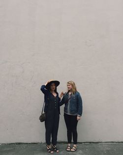 Full length of female friends standing on sidewalk against beige wall