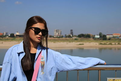 Full length of young woman wearing sunglasses standing by railing against sky