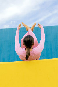 Back view of unrecognizable female in light pink activewear lying on back with knees to chest with leg crossed while doing yoga exercises on blue and yellow background