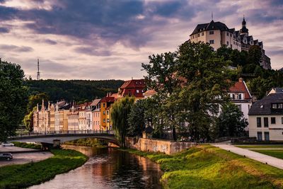 Castle and buildings by river against sky in city