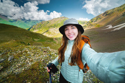 Happy young woman tourist smiling making selfie on phone against mountains and fluffy clouds, hike