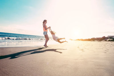 Full length of playful mother spinning daughter while holding her hands at beach during sunny day