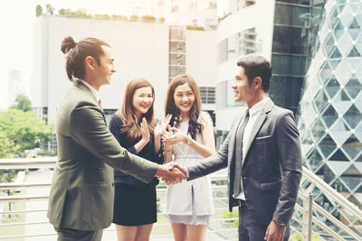 Smiling colleagues clapping while businessmen handshaking against buildings in city