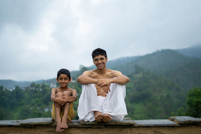 A young man sitting on the roof with his younger brother,both wearing dhoti, smiling into the camera