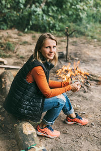 A young woman is warming herself by the fire with a cup of warming tea in the forest