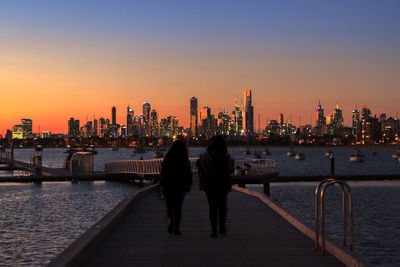 Friends walking on pier over sea against buildings during sunset