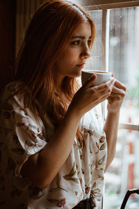 Thoughtful young woman looking through window while drinking coffee in cup at home