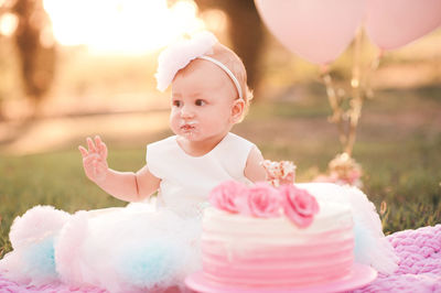 Baby girl 1 year old eating creamy birthday cake sitting on green grass with pink balloons outdoor