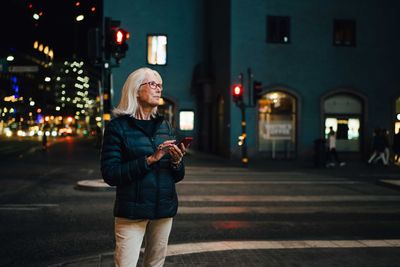 Smiling woman using smart phone while looking up in illuminated city at night