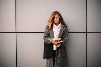 Young woman using phone while standing against wall