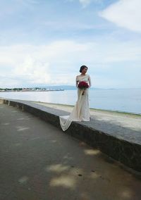 Full length of woman holding flower bouquet by sea against sky