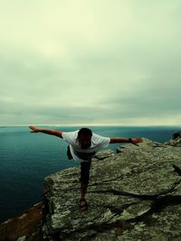 Full length of man standing on one leg over rock against sea and sky