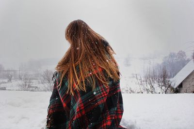 Woman wrapped in blanket sitting on snow covered landscape against sky